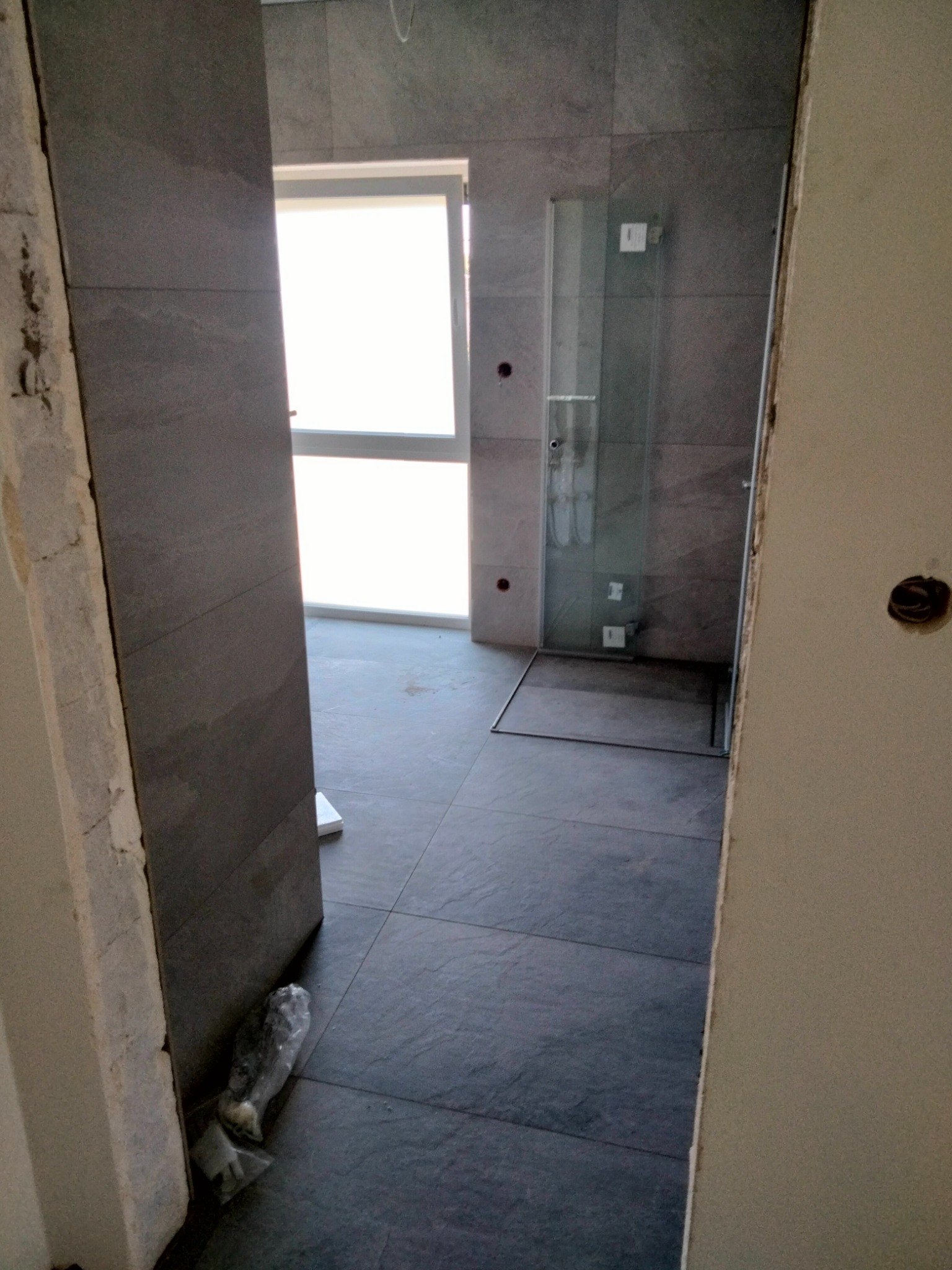 Wnętrze łazienki w trakcie prac wykończeniowych, widoczne szare płytki na ścianach i podłodze, zamontowana kabina prysznicowa i okno z widokiem na zewnątrz, fragment ściany z surowym wykończeniem...