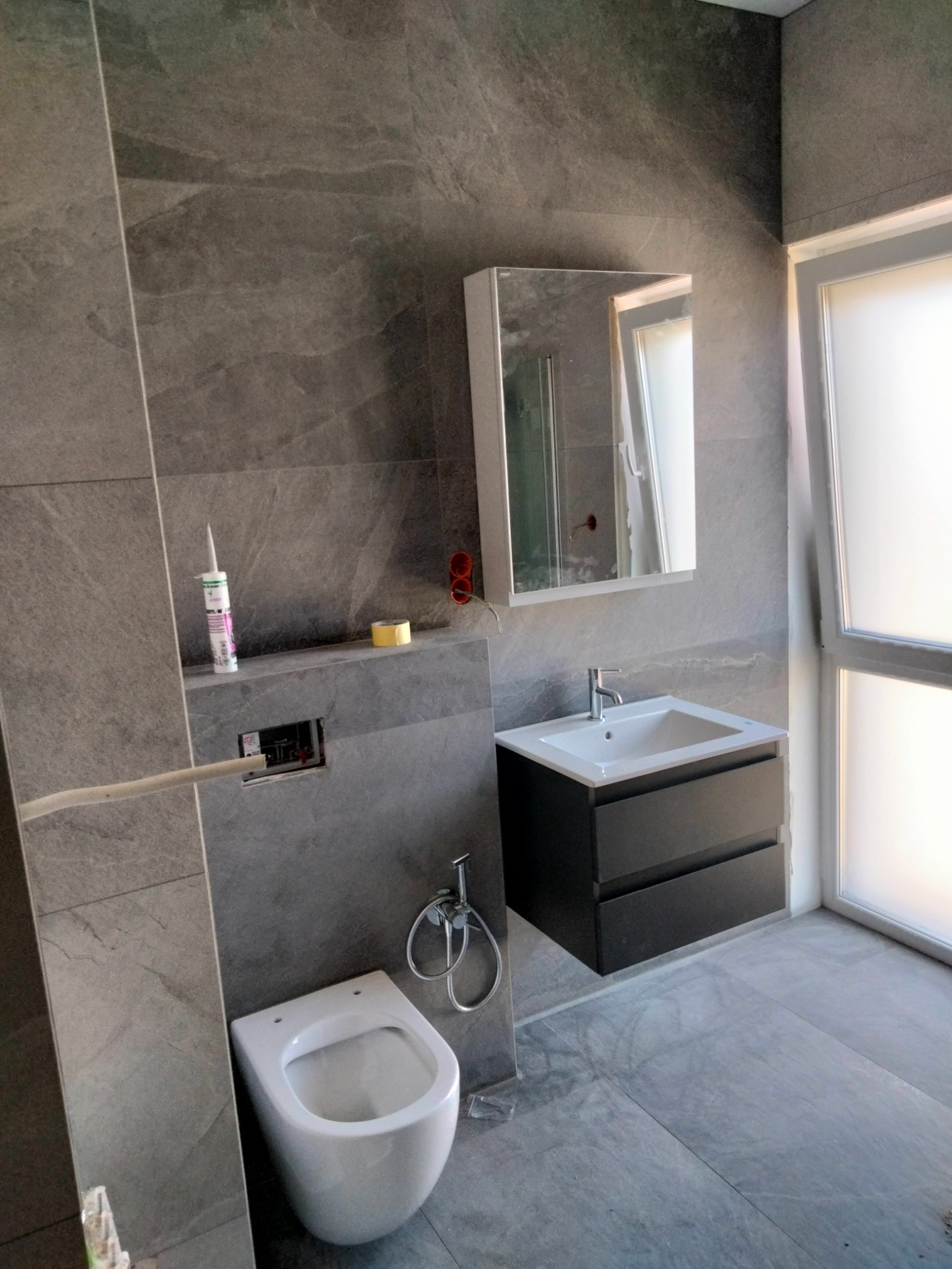 Szara łazienka w trakcie wykańczania z podwieszaną toaletą, szafką z umywalką i lustrem, widoczne materiały budowlane i niedokończone instalacje.