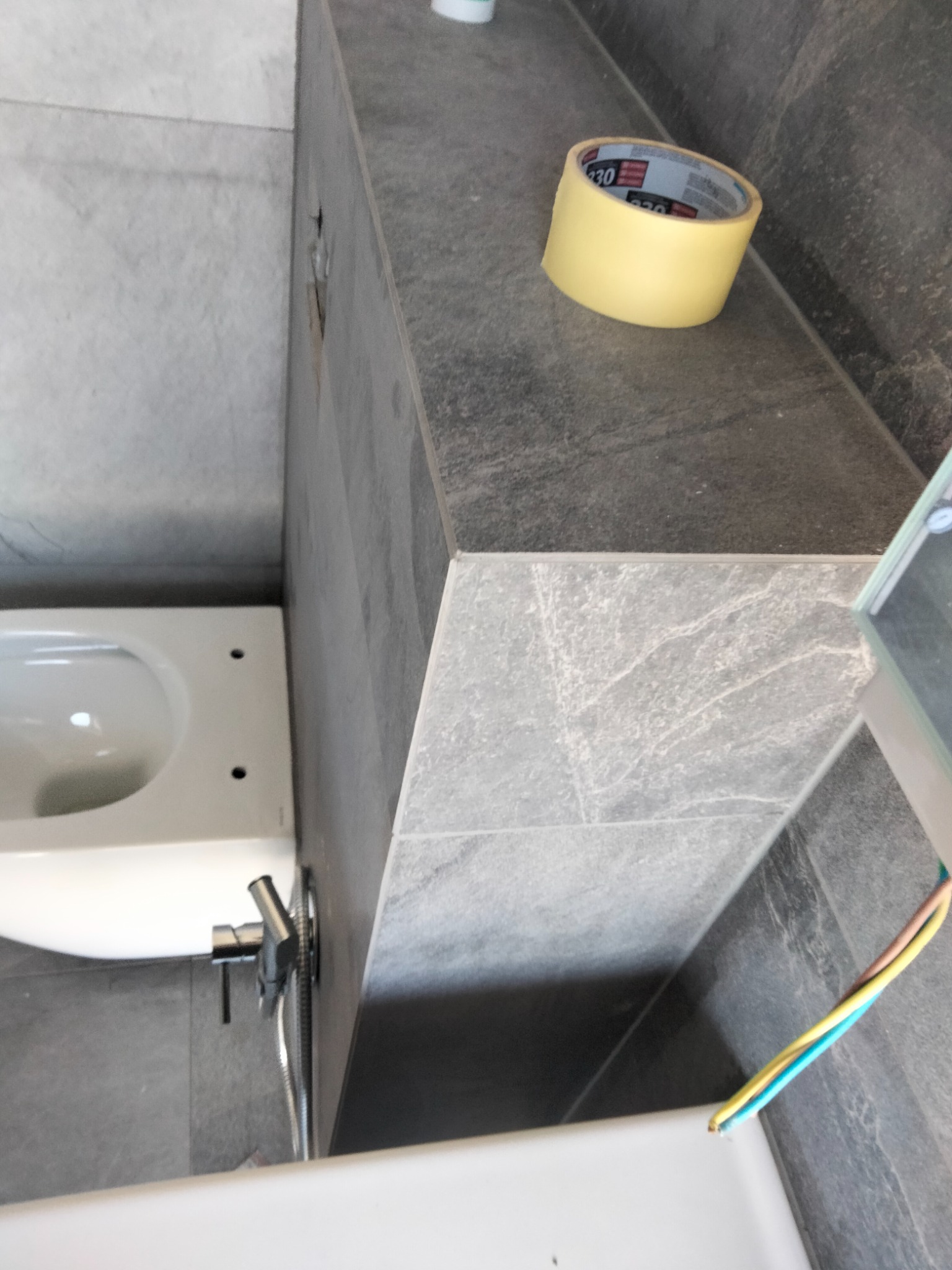 Fragment łazienki w trakcie remontu z widoczną białą miską WC, szarą glazurą imitującą kamień, taśmą malarską na blacie i wystającymi przewodami elektrycznymi.