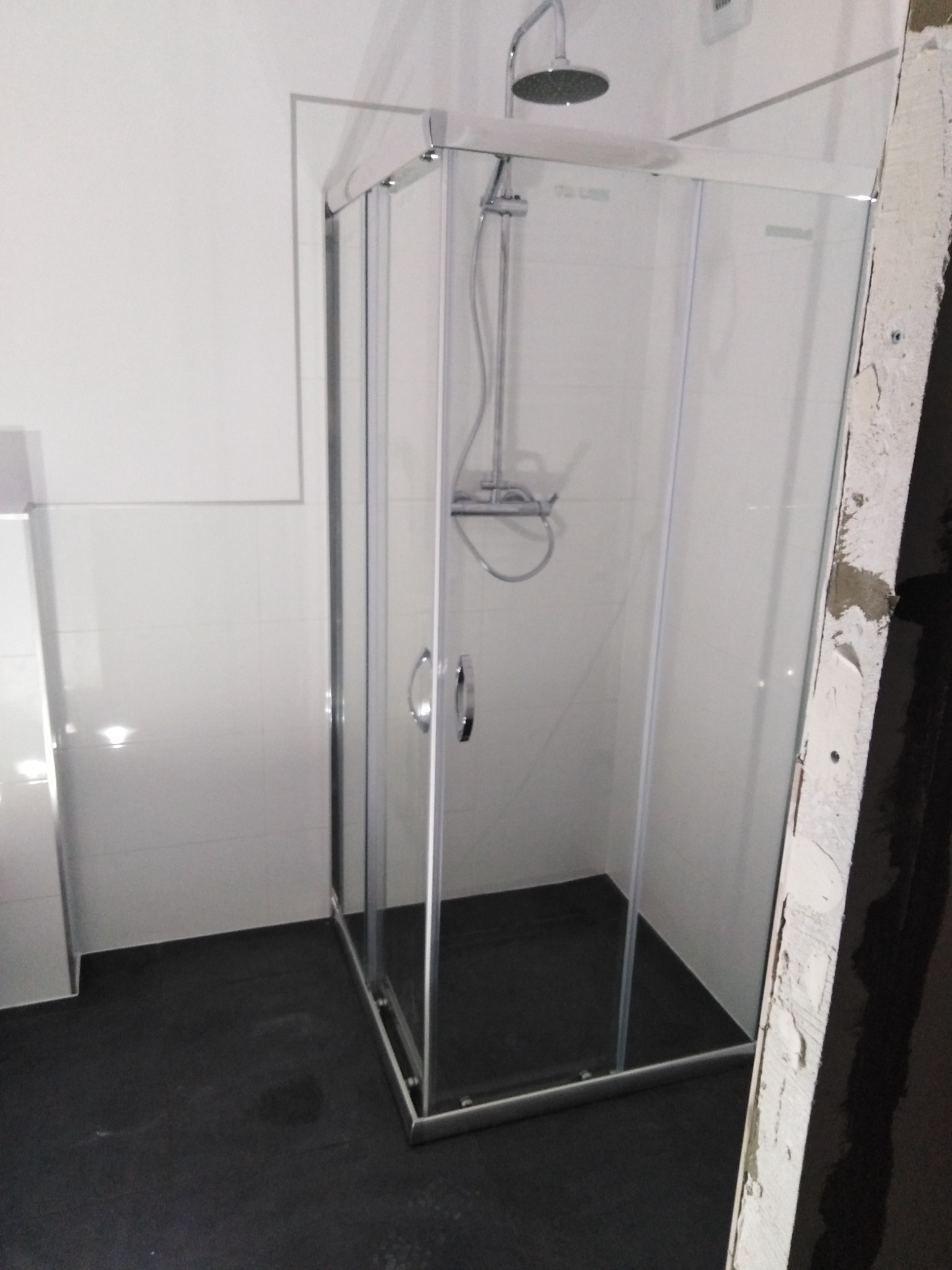 Nowoczesna kabina prysznicowa z chromowanymi profilami i deszczownicą, zamontowana w wykończonej łazience z ciemną podłogą i białymi płytkami ściennymi, widoczny fragment ściany w trakcie prac...
