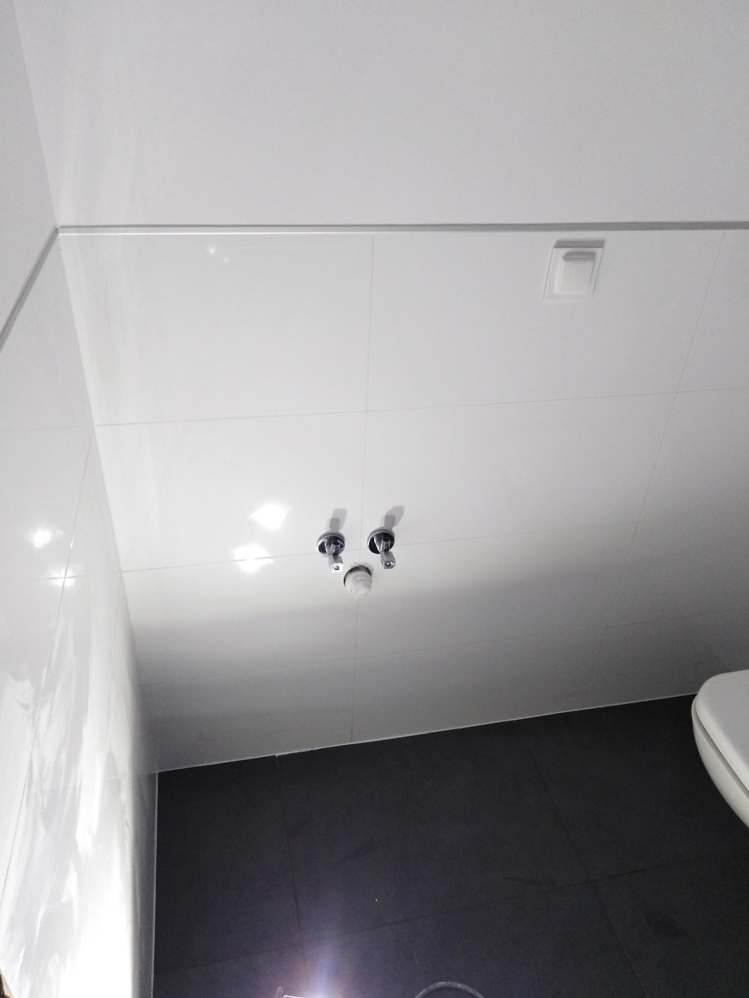 Nowoczesna łazienka z białymi płytkami na ścianach i suficie, ciemnoszarą podłogą i widocznymi przyłączami wodnymi do baterii umywalkowej.