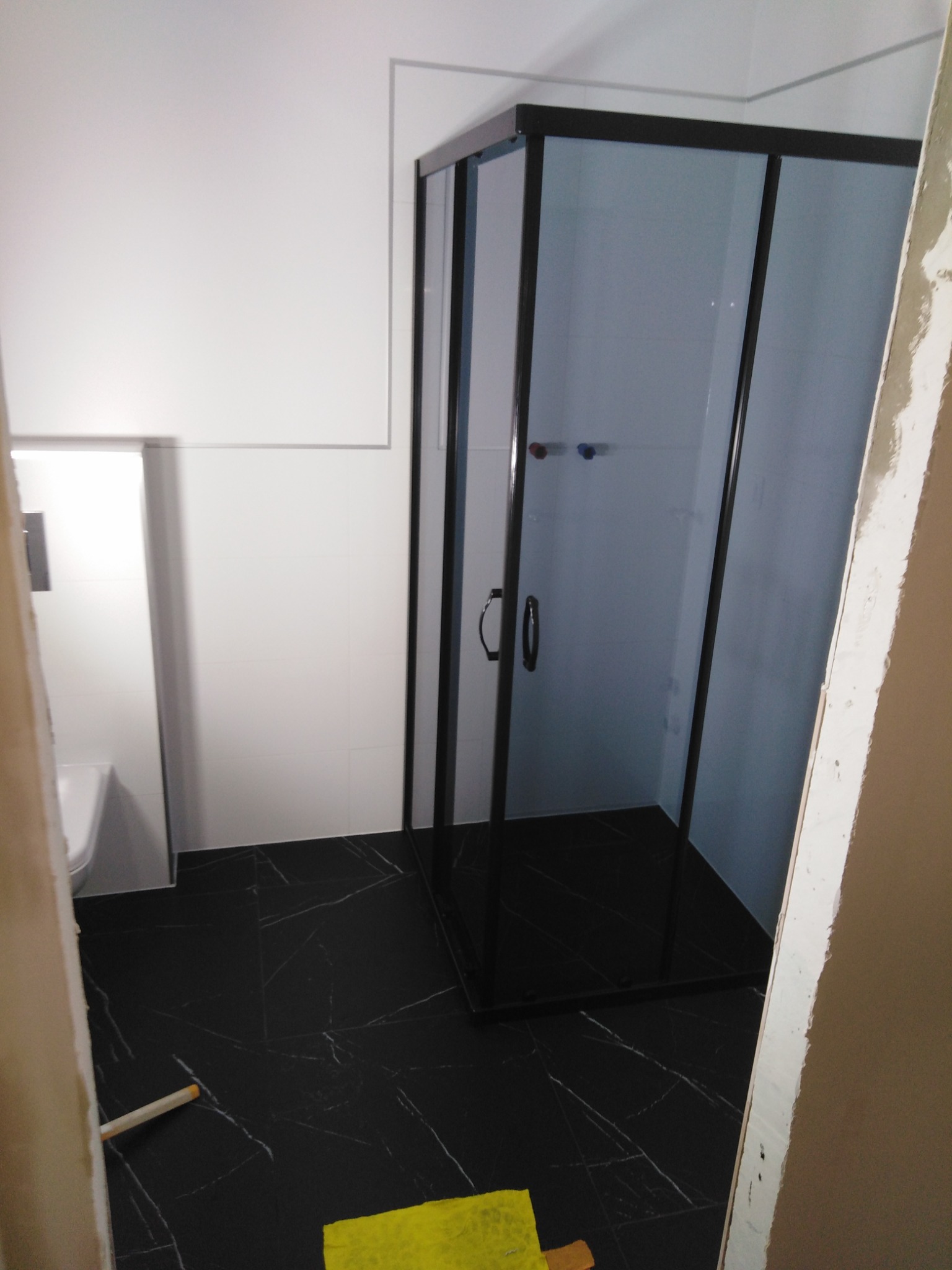 Częściowo wykończona łazienka z czarną kabiną prysznicową z czarnymi profilami, czarną marmurkową podłogą, białą toaletą podwieszaną i fragmentem niezakończonej ściany z widocznymi śladami gładzi.