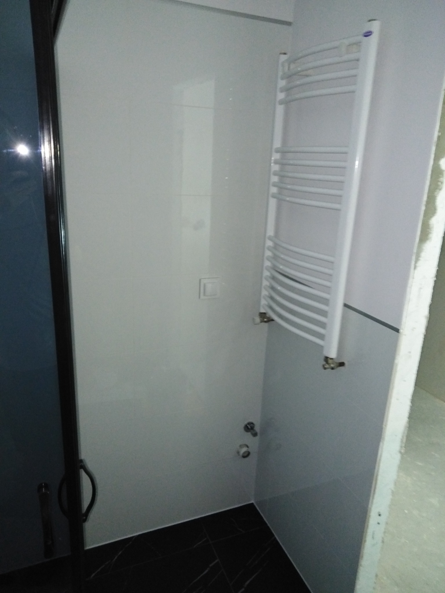 Fragment łazienki z kabiną prysznicową z ciemnymi drzwiami, białymi płytkami na ścianach, grzejnikiem drabinkowym i czarną podłogą z białymi żyłkami.