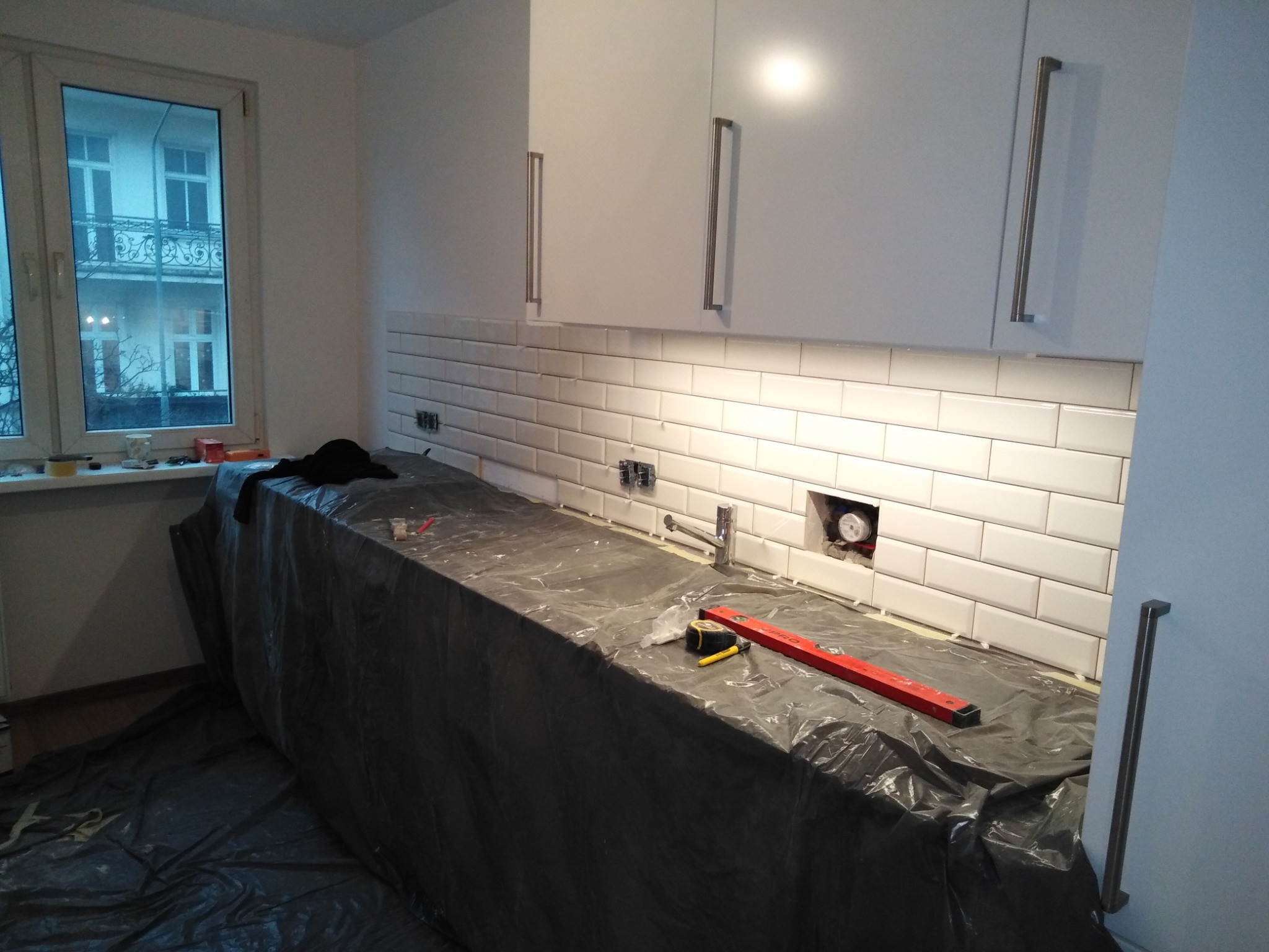 Białe, prostokątne płytki ułożone na ścianie w kuchni, widoczna instalacja wodna, blat zabezpieczony folią, poziomica i miarka leżą na blacie, białe szafki wiszące z długimi, srebrnymi uchwytami.