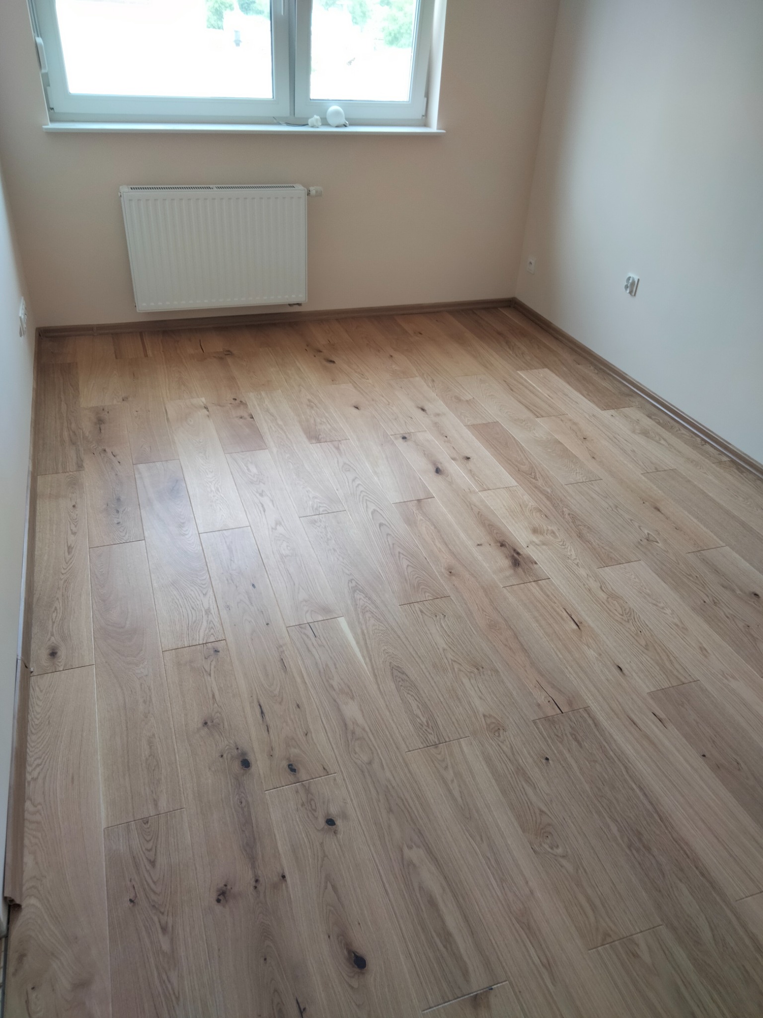 Jasne, świeżo ułożone deski parkietowe z widocznymi naturalnymi słojami i sękami, wypełniające wnętrze pokoju z białymi ścianami, grzejnikiem i oknem.