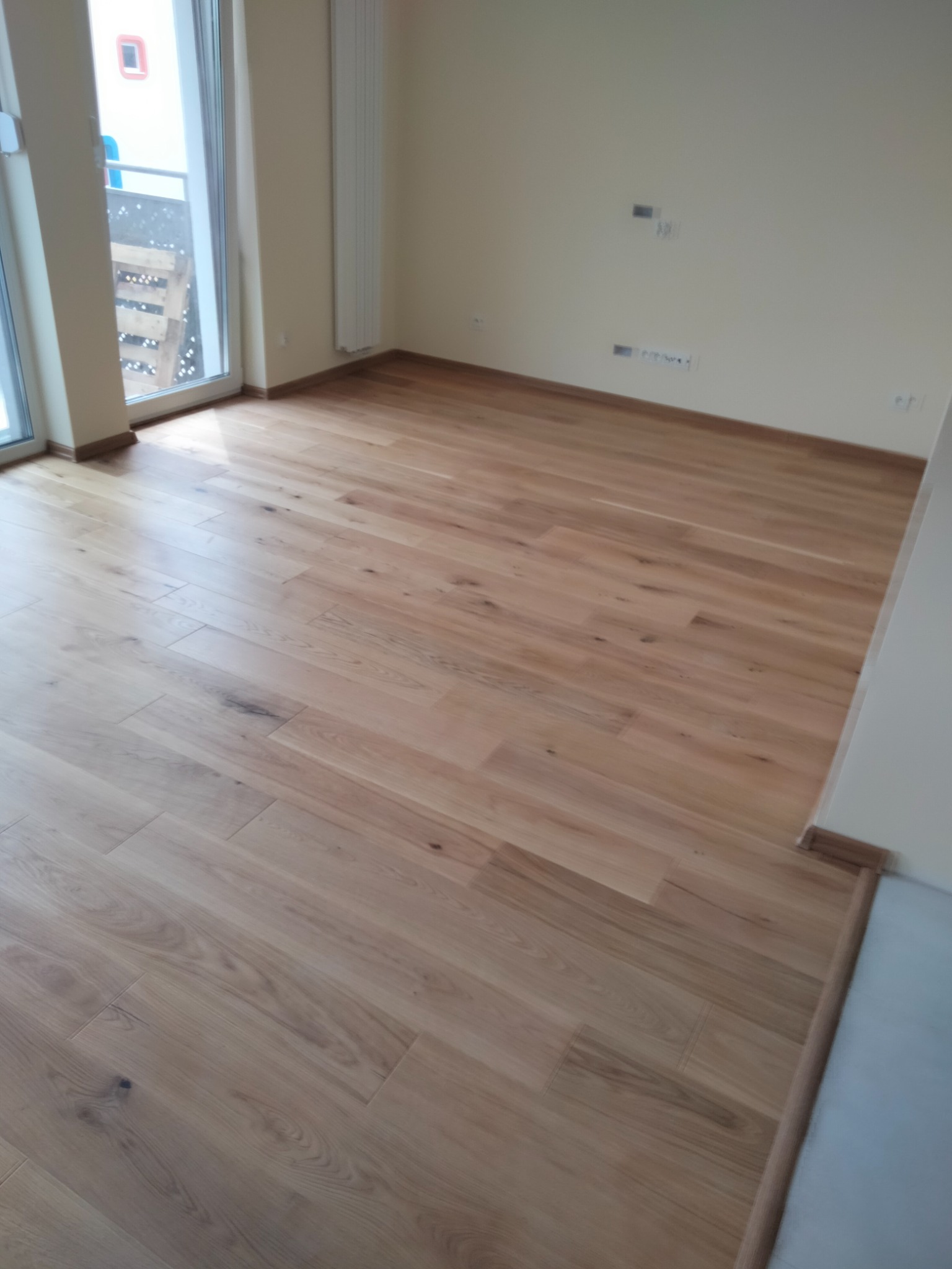 Jasny, drewniany parkiet ułożony w pustym pokoju z widokiem na balkon przez duże okno, widoczne listwy przypodłogowe i grzejnik na ścianie.