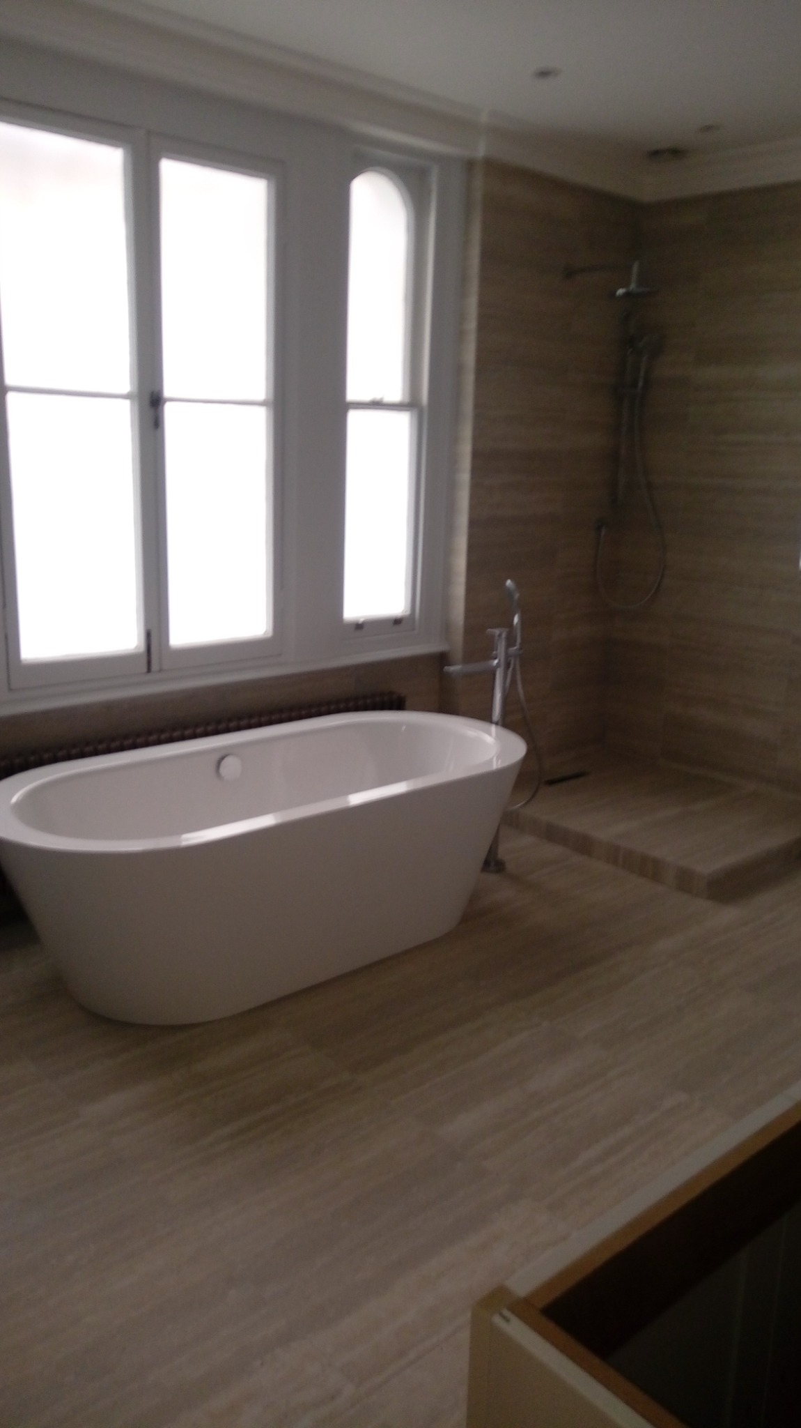 Nowoczesna łazienka z wolnostojącą wanną przy oknie, wykończona płytkami imitującymi drewno i kabiną prysznicową bez brodzika.