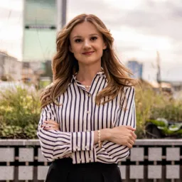 Uśmiechnięta kobieta w bluzce w paski, z założonymi rękami, pozuje na tle miejskiego krajobrazu z zielenią.