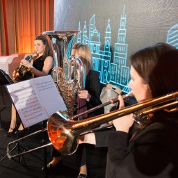 Warsaw Womens Brass Quintet - Kwartet instrumentalny grający na scenie z podświetlanym tłem przedstawiającym abstrakcyjny pejzaż miejski w neonowych barwach.