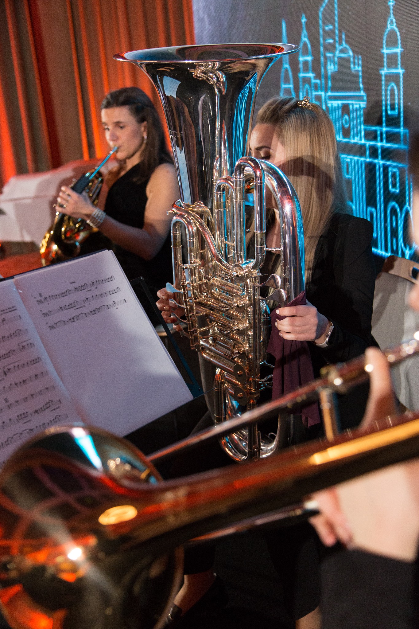 Kobieta stroi tubę na scenie, obok nuty i inni muzycy z instrumentami dętymi, w tle dekoracja świetlna zarysów miasta.