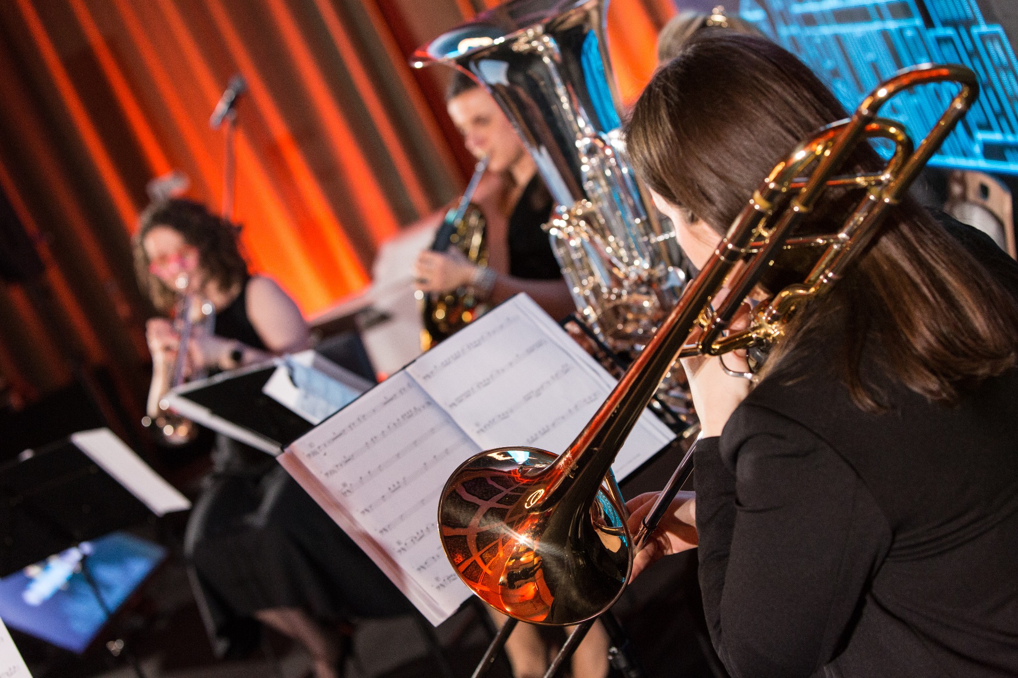 Orkiestra dęta na scenie: puzonistka z tyłu, w tle trąbka i tuba, nuty na pulpicie, pomarańczowe i niebieskie oświetlenie sceniczne.