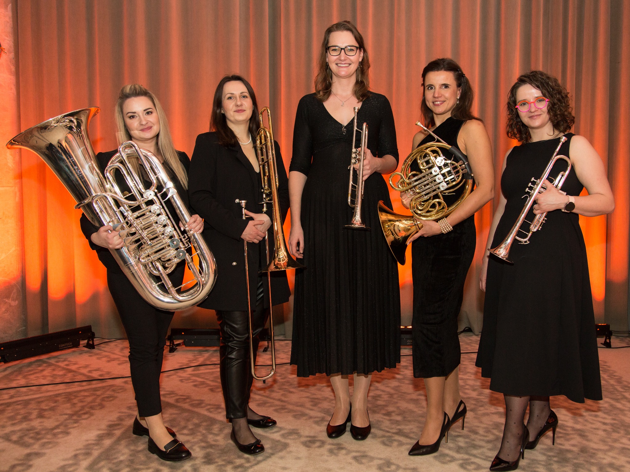 Pięć kobiet w czarnych sukienkach i eleganckich butach pozuje z instrumentami dętymi: tubą, puzonem, trąbką i waltornią na tle pomarańczowej zasłony.