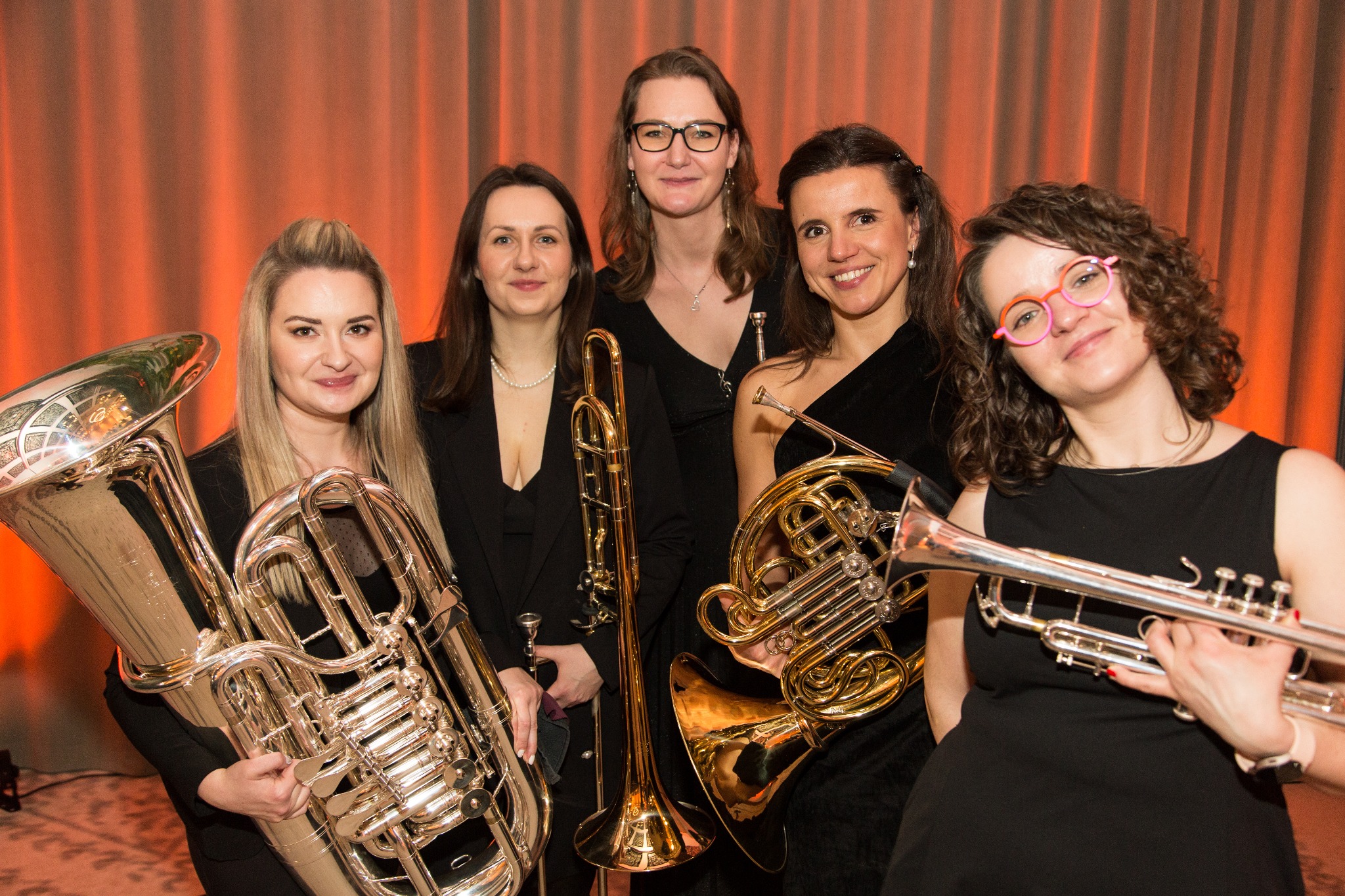 Kobiecy kwintet dęty w czarnych strojach pozuje z instrumentami: tuba, puzon, róg i trąbka na tle pomarańczowej zasłony.