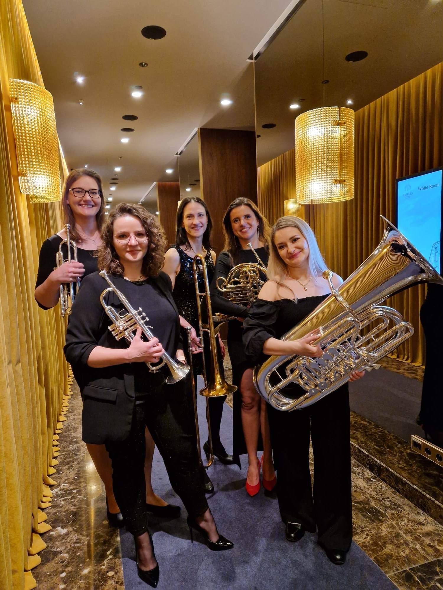 Pięć uśmiechniętych kobiet w eleganckich, czarnych strojach pozuje z instrumentami dętymi: trąbkami, puzonem, waltornią i tubą, w korytarzu z dekoracyjnymi, złotymi zasłonami i lampami.