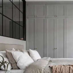 Szara zabudowa szafy z frezowanymi drzwiami i złotymi gałkami, widoczna za łóżkiem z jasną pościelą i dekoracyjną gałązką eukaliptusa, obok czarna rama okna z podziałami.