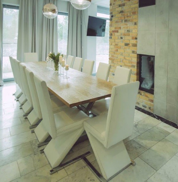 Jadalnia z długim drewnianym stołem otoczonym białymi krzesłami na metalowych nogach, kominek z cegły i płytek w tle, widoczne lampy wiszące i okno z zasłonami.