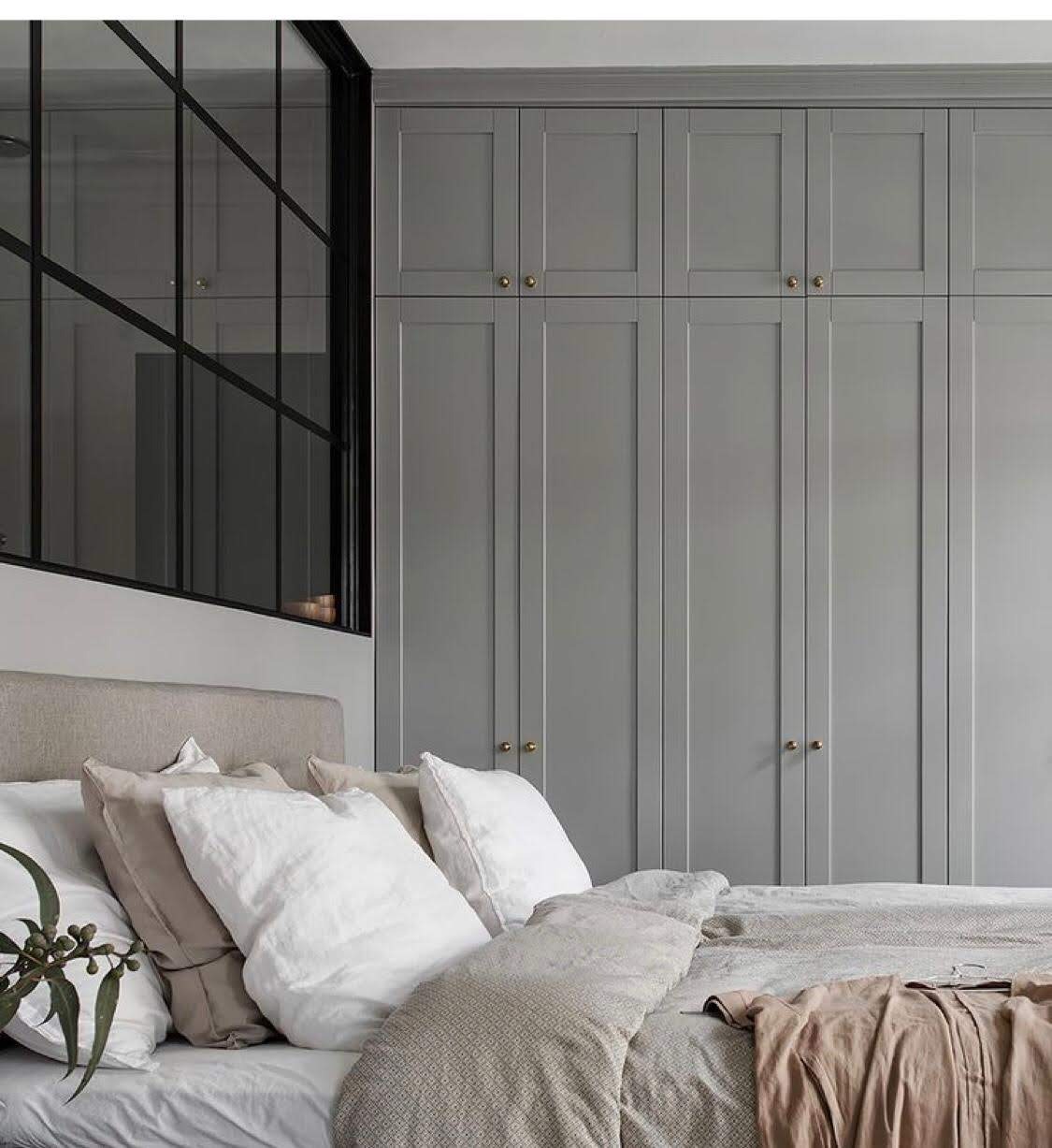 Szara zabudowa szafy z frezowanymi drzwiami i złotymi gałkami, widoczna za łóżkiem z jasną pościelą i dekoracyjną gałązką eukaliptusa, obok czarna rama okna z podziałami.