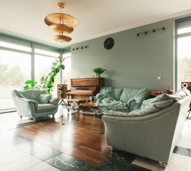 Jasny salon z parkietem, pastelowymi meblami, pianinem, roślinami i dekoracyjnym oświetleniem sufitowym, widok na zewnątrz przez duże okna.