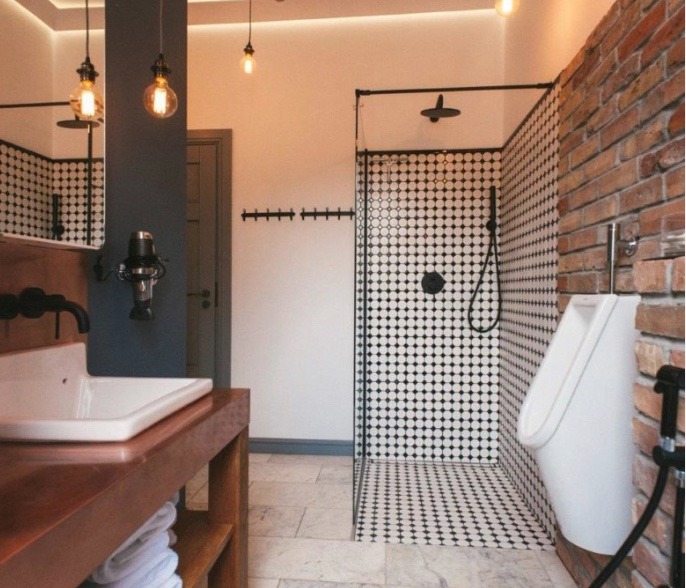 Łazienka w stylu industrialnym z kabiną prysznicową wyłożoną czarno-białą mozaiką, umywalką na drewnianym blacie, pisuar na tle ceglanej ściany i żarówkami Edisona.