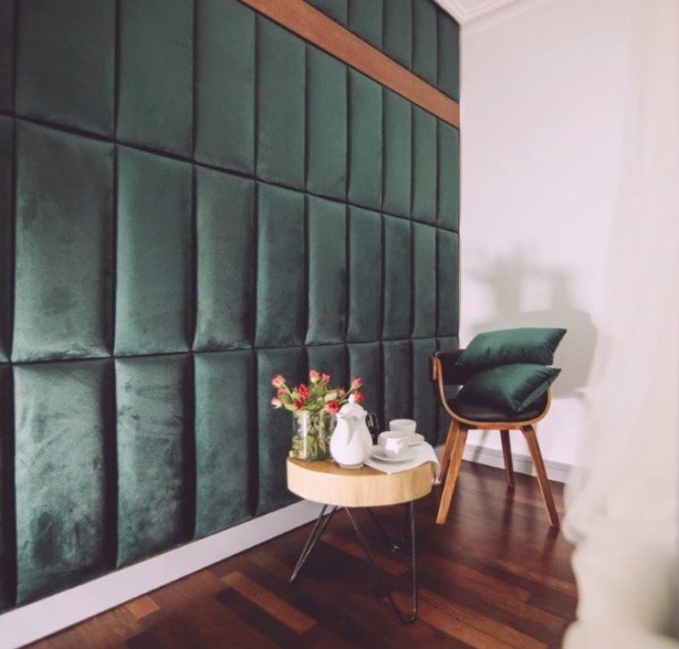 Ściana wykończona ciemnozielonymi panelami tapicerowanymi z drewnianym akcentem, stolik kawowy z kwiatami i serwisem do kawy oraz krzesło z poduszkami w rogu pokoju.