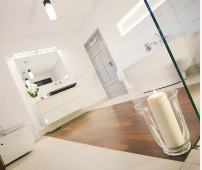 Jasna łazienka z drewnianą podłogą, białą wanną na nóżkach, umywalką z szafkami i zapaloną świecą w szklanym świeczniku na pierwszym planie.