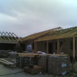 Budowa domu w trakcie: mury z cegły, drewniana konstrukcja dachu, palety z materiałami budowlanymi na placu budowy, pochmurne niebo.