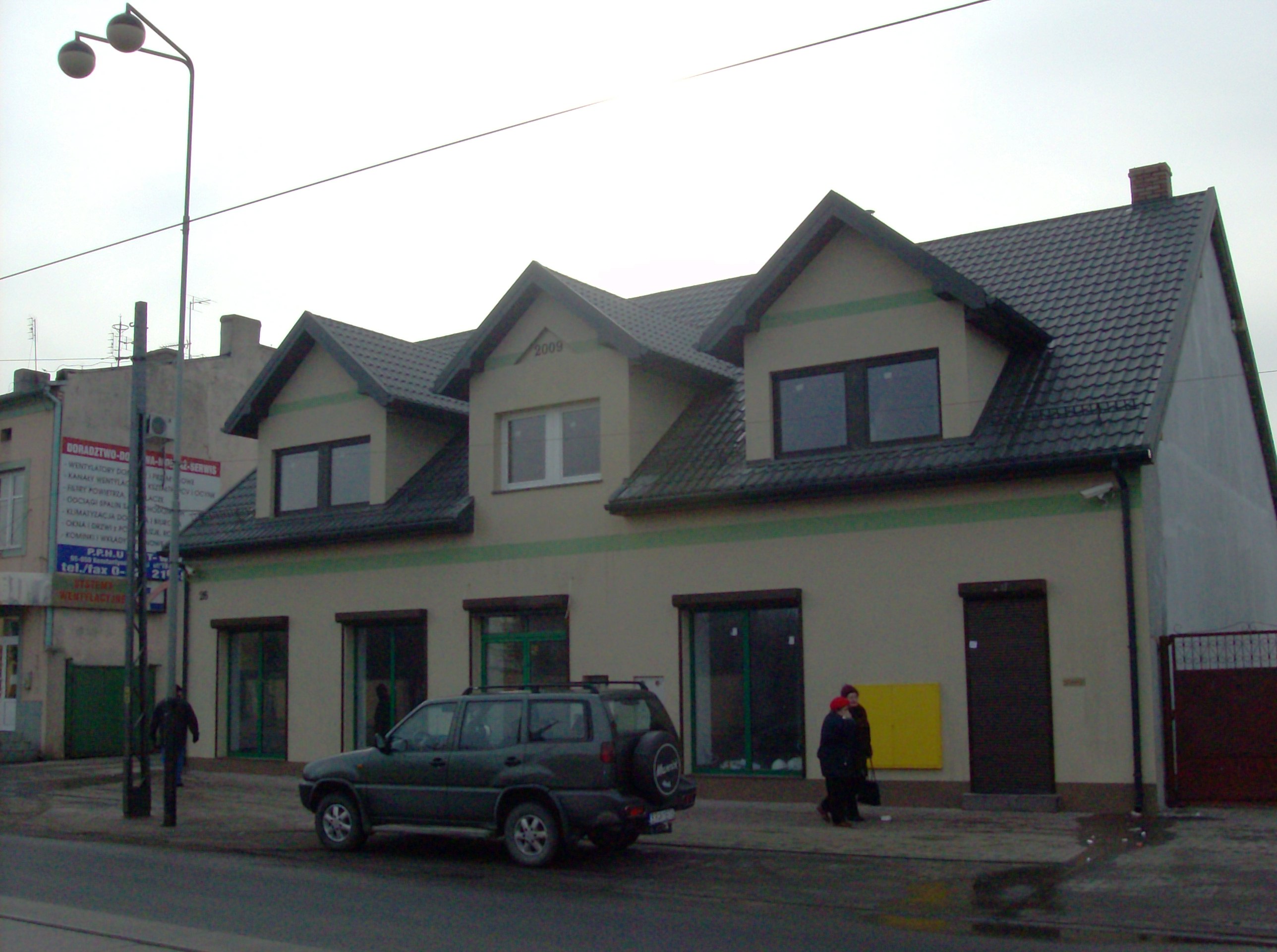 Budynek mieszkalny z poddaszem użytkowym, pomalowany na beżowo z ciemnymi ramami okiennymi i dachem z ciemnej dachówki, rok budowy 2009, przed budynkiem zaparkowany samochód terenowy, na chodniku...