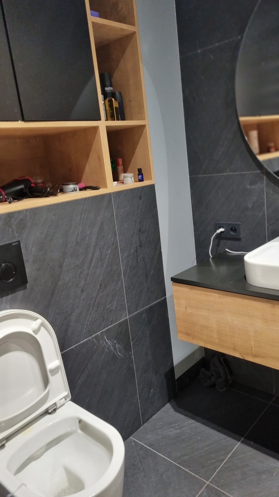 Nowoczesna łazienka z ciemnymi płytkami imitującymi kamień, drewnianymi półkami i szafką pod umywalką; widoczna toaleta z otwartą klapą, okrągłe lustro i akcesoria na półkach.