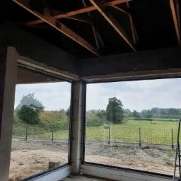 Surowy stan wnętrza z zamontowanym narożnym oknem z czarną ramą, widok na łąkę z belami siana przez szybę, widoczna konstrukcja dachu.