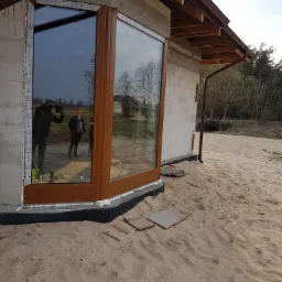 Montaż brązowej ramy okiennej w surowym budynku z betonu komórkowego, widoczne narzędzia i odbicie dwóch osób w szybie.