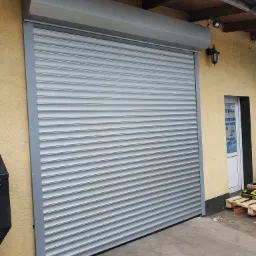 Zamknięta, szara roleta zewnętrzna na budynku o żółtej elewacji, widoczna boczna lampa i drzwi z reklamą na szybie.