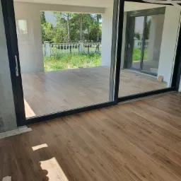 Duże przesuwne drzwi balkonowe z czarną ramą, widok na taras z balustradą i zieleń, drewnopodobna podłoga wewnątrz.