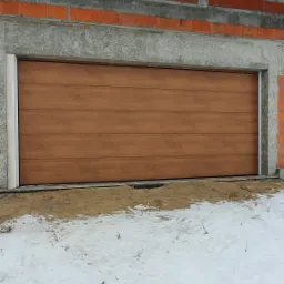 Zamknięta, segmentowa brama garażowa w kolorze drewna, zamontowana w surowym stanie budynku z widocznymi cegłami i betonem, przed bramą śnieg.