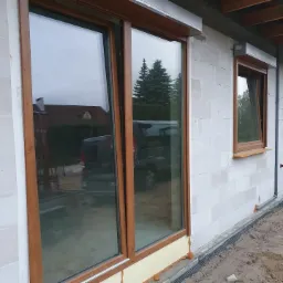 Montaż okien PCV z brązowymi ramami i szarymi roletami zewnętrznymi w surowym stanie budynku z białego bloczka.