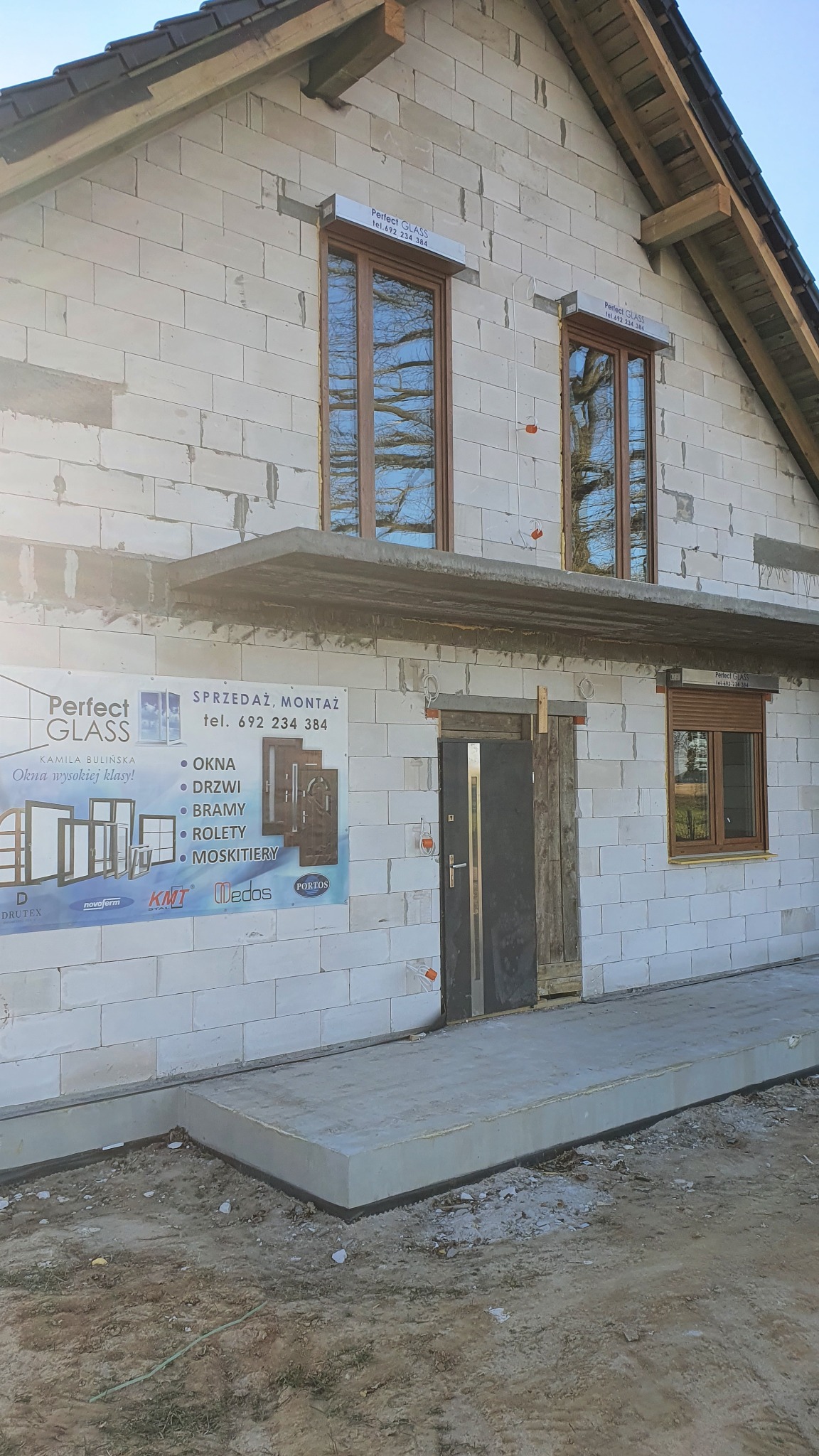 Wznoszony budynek z zamontowanymi oknami z brązowymi ramami, częściowo zasłoniętą roletą i reklamą firmy na ścianie.