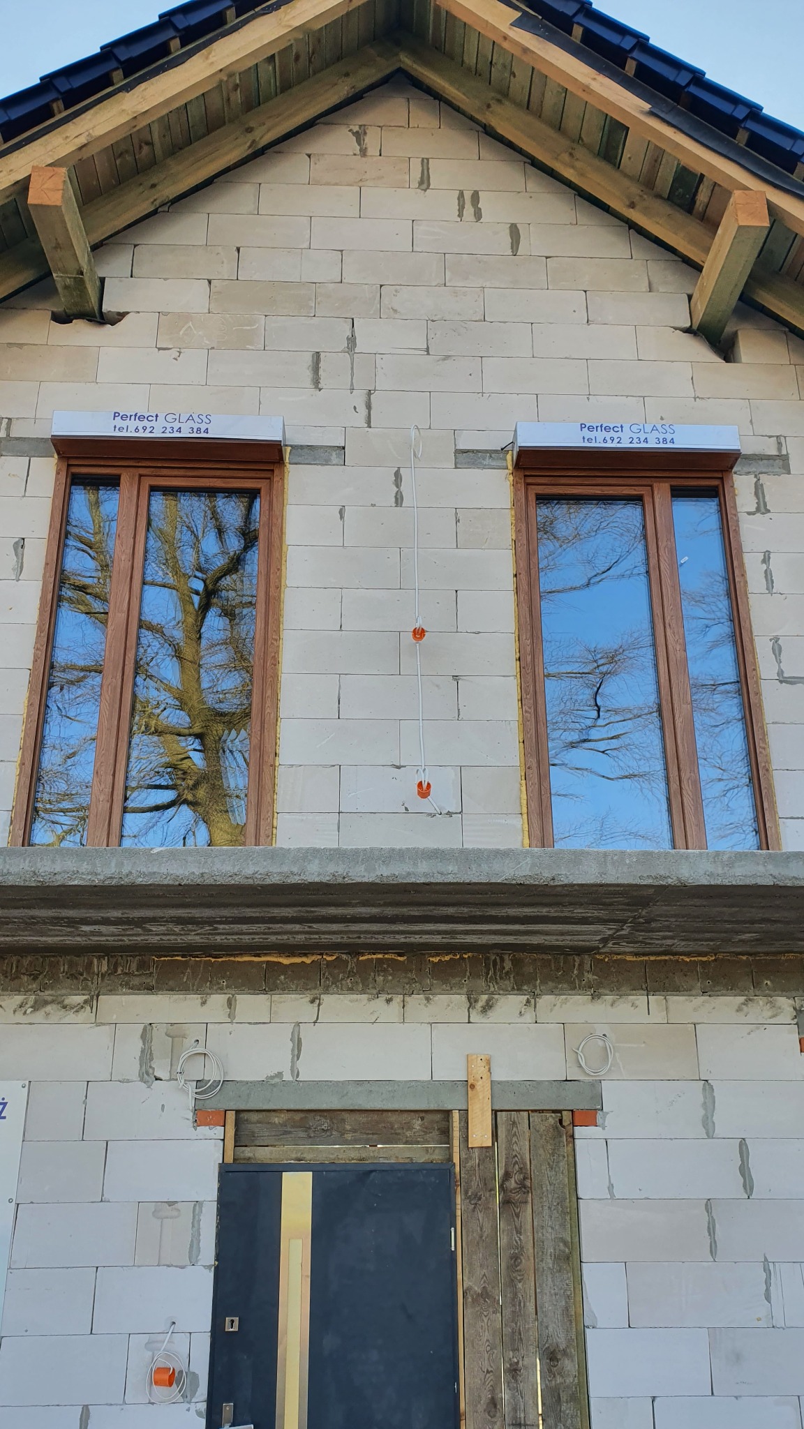 Surowy stan budynku w trakcie budowy z dwoma oknami z brązowymi ramami, w których odbija się drzewo i niebieskie niebo. Nad oknami widoczne zwinięte rolety z napisem 'Perfect Glass'.