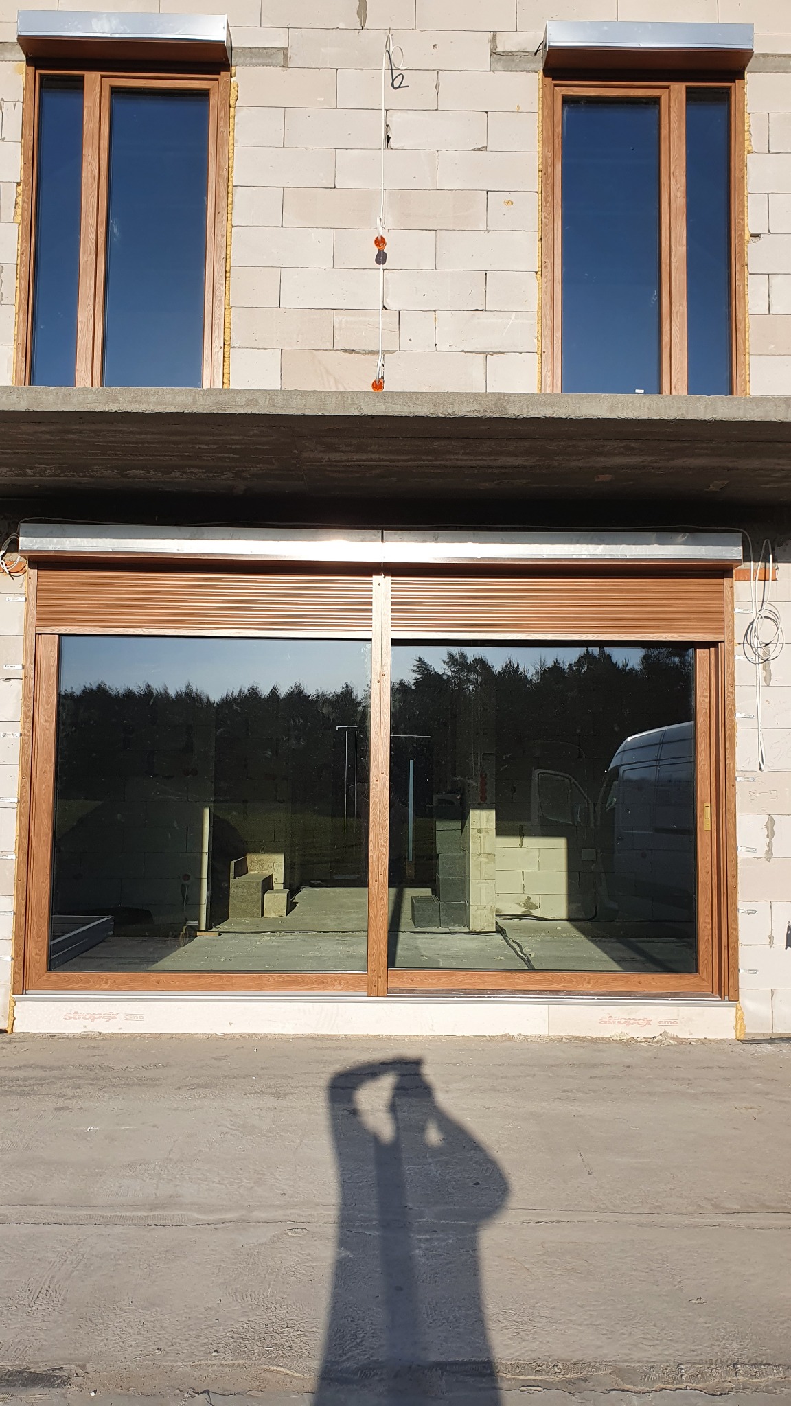 Montaż okien z brązowymi ramami i zewnętrzną roletą w surowym stanie budynku, widoczny cień osoby robiącej zdjęcie.