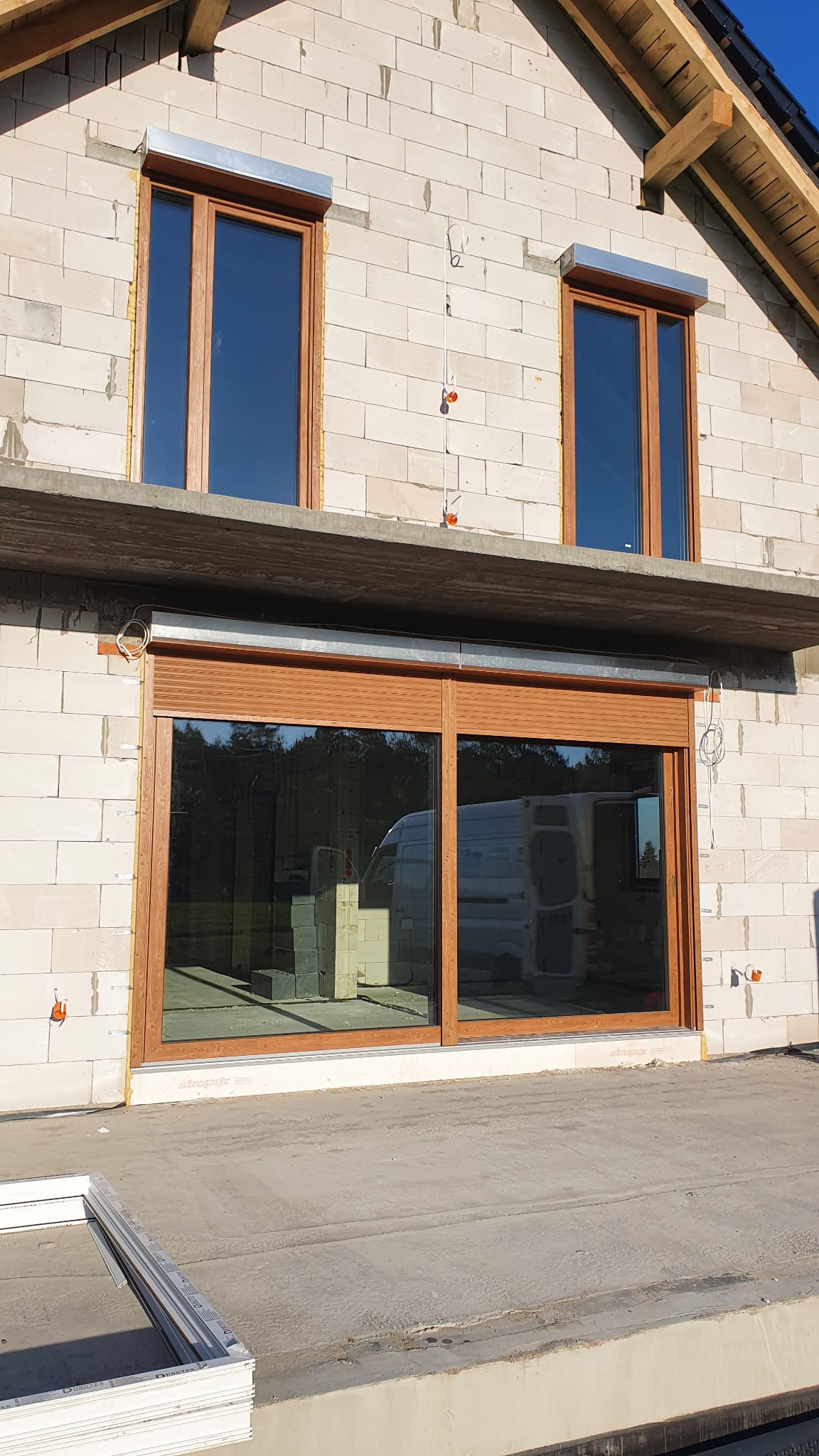 Surowy stan budynku z zamontowanymi oknami z brązowymi ramami i zewnętrznymi roletami w kolorze drewna, widoczne elementy instalacji elektrycznej i białe bloczki konstrukcyjne, na dole okna...