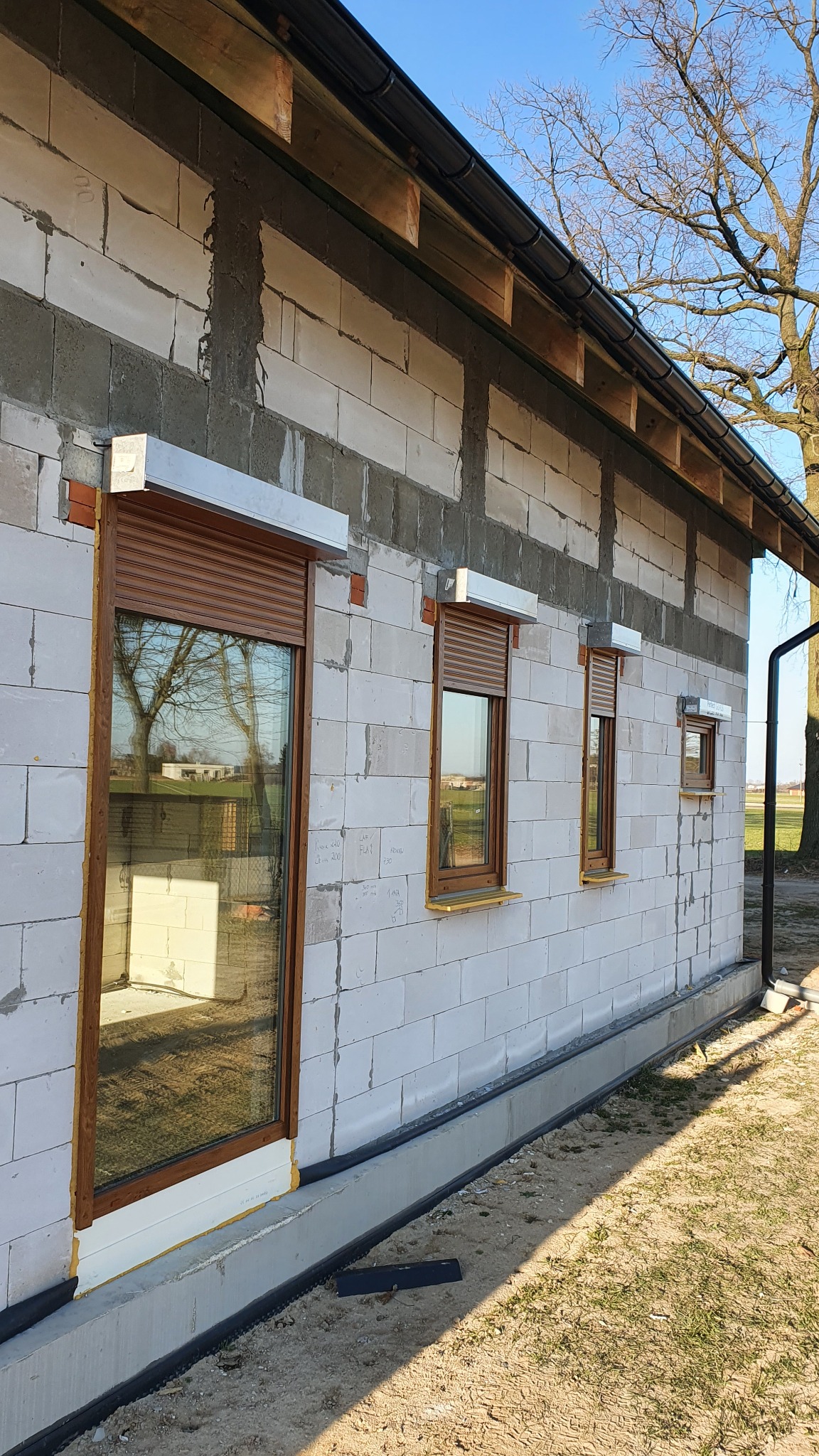 Surowy budynek z białych bloczków z zamontowanymi oknami z brązowymi roletami zewnętrznymi, widoczne detale montażu.