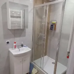 Nowoczesna łazienka po remoncie: kabina prysznicowa z chromowaną armaturą, biała umywalka z szafką, lustro z grzejnikiem drabinkowym, szare płytki ścienne i podłogowe imitujące drewno.