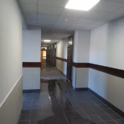 Korytarz w budynku z świeżo położonymi, ciemnymi płytkami podłogowymi, częściowo mokrymi, odblaskującymi światło, z jasnymi ścianami wykończonymi do połowy wysokości jasnymi płytkami, zakończonymi...