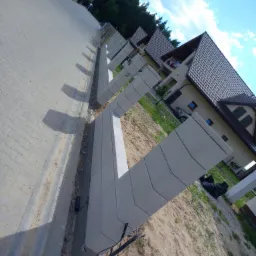 Nowoczesny mur oporowy z prefabrykatów betonowych w trakcie budowy, widoczne kable i przewody, w tle dom jednorodzinny i chodnik z kostki brukowej.