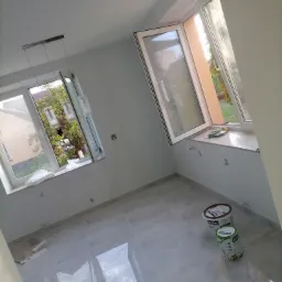 Pomalowane na jasno szaro pomieszczenie z dwoma oknami, na podłodze płytki imitujące marmur, widoczne dwa wiadra z farbą Śnieżka.