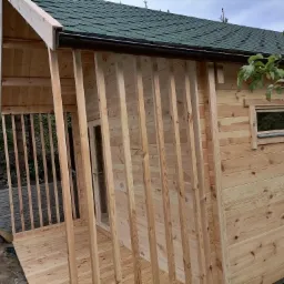 Drewniana konstrukcja domku letniskowego w trakcie budowy, widoczne deski elewacyjne, zielony dach z gontu bitumicznego, montaż rynien i częściowo wykonana weranda z drewnianymi słupkami.