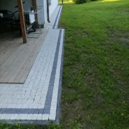 Ułożona kostka brukowa w odcieniach szarości z ciemniejszym pasem obok drewnianego tarasu i trawnika, widoczny fragment elewacji budynku z rynną.