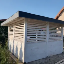 Biała, drewniana altana z czarnym, płaskim dachem, otoczona tujami, w tle dom z brązowym dachem i fragment białego samochodu dostawczego.