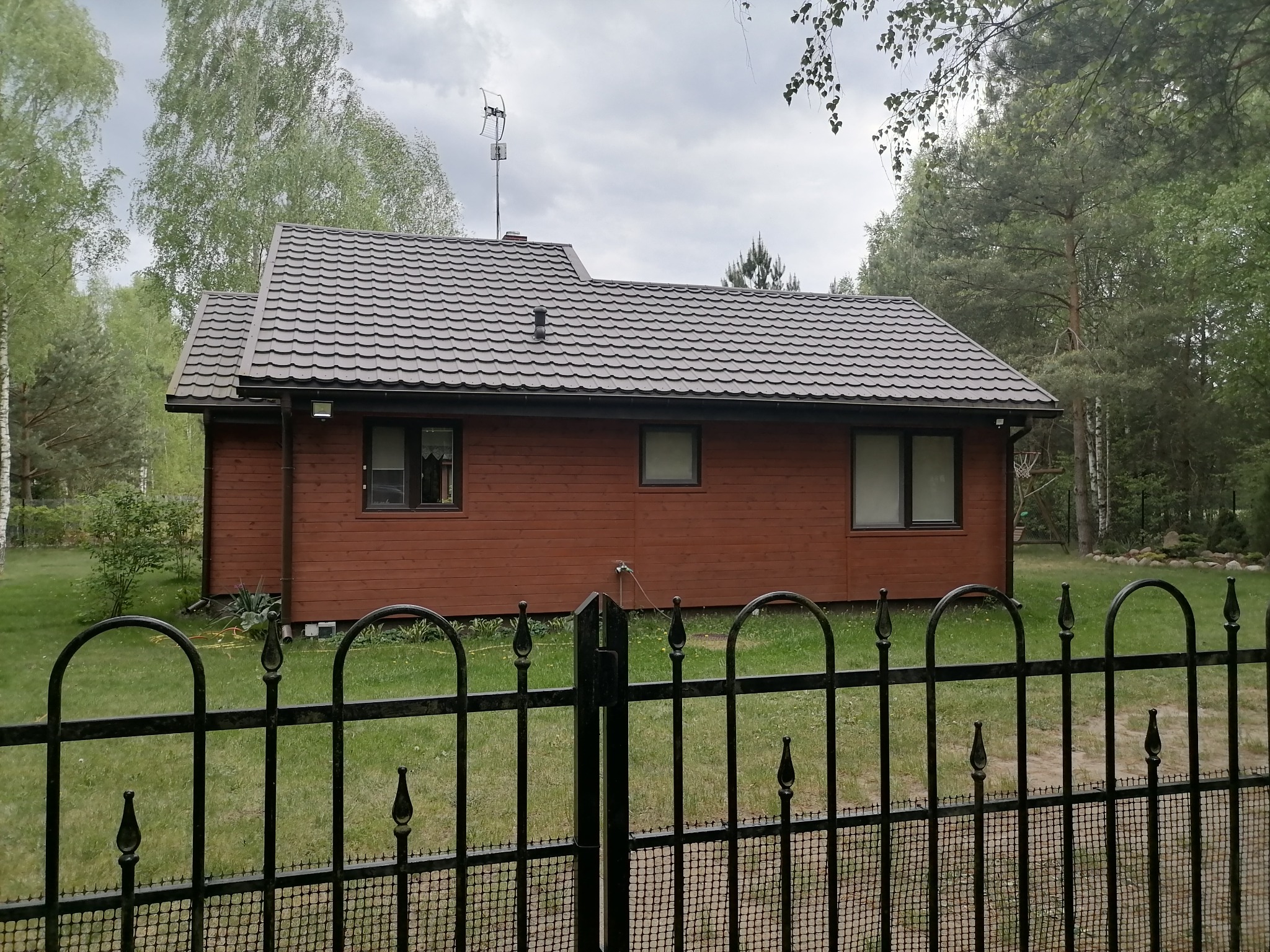 Dom z brązową drewnianą elewacją i ciemnoszarym dachem z blachodachówki, widoczny zza metalowego ogrodzenia w pochmurny dzień.
