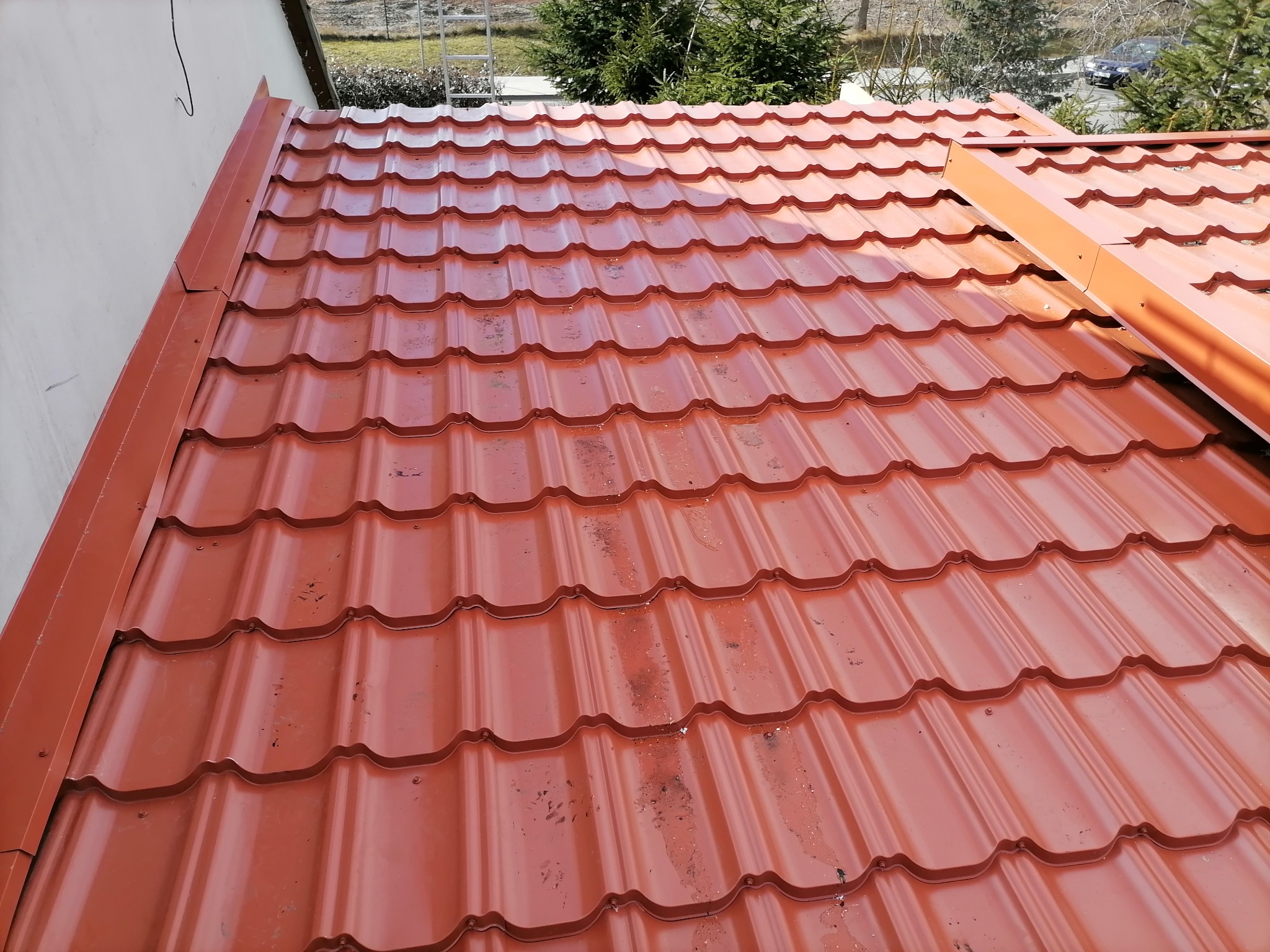 Czerwona blachodachówka z zamontowanymi rynnami, widoczne ślady zabrudzeń na powierzchni dachu, w tle drzewa iglaste.