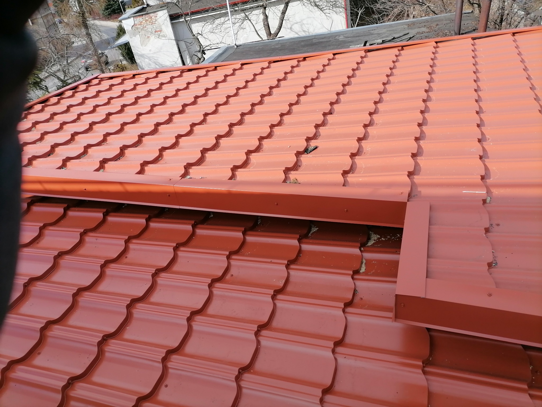 Czerwona blachodachówka ułożona na dachu budynku, widoczne elementy orynnowania, fragment dachu sąsiedniego budynku i drzewa w tle.