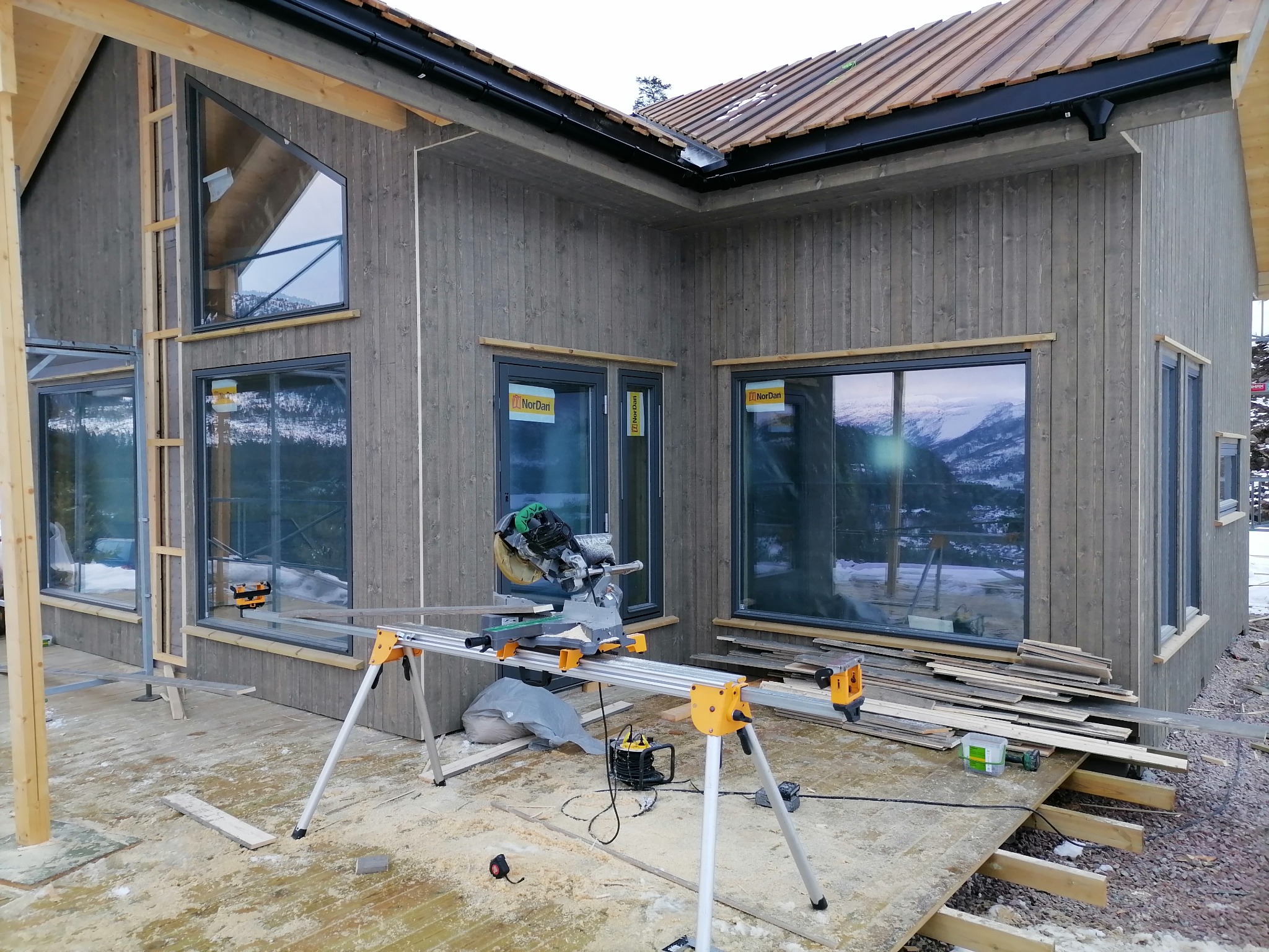 Szary dom z drewnianą elewacją w trakcie budowy, widoczna piła stołowa, okna z widokiem na góry i częściowo położony dach z brązowej blachy.